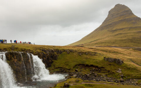 South Greenland Exploration cruise travelers stand atop the Kirkjufellsfoss waterfall near a grassy peak on a cloudy day in Iceland.
