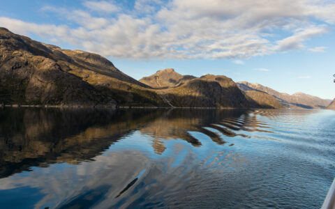 View from Nat Geo Endurance ship as she passes through Kuannersooq Fjord in Greenland with glassy water & rolling rocky hills.