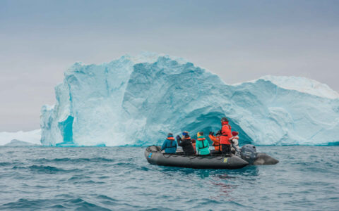 Zodiac boat with south Greenland Exploration cruise guests in colorful coats sits in front of large blue iceberg in calm sea.