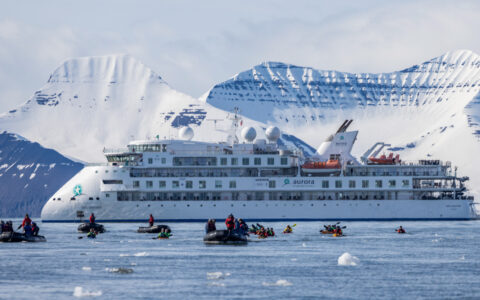 White expedition ship Greg Mortimer sits in calm icy water by snow-covered peaks as travelers kayak & Zodiac cruise nearby.
