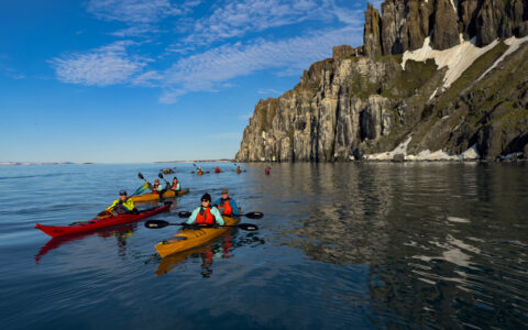 Tandem kayakers in red & yellow boats sit atop glassy water beside tall, rocky cliffs on a sunny day in Spitsbergen.