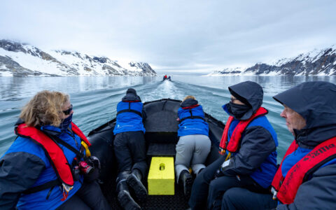 Spitsbergen: Realm of the Ice Bear Cruise guests in red life jackets & blue coats sit in Zodiac & cruise among snowy peaks.