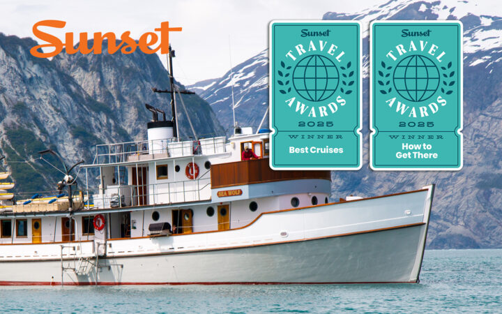 Small wooden ship Sea Wolf in Glacier Bay National Park Alaska surrounded by mountain range. Sunset magazine logo and two travel awards badges