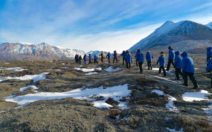 Group of Arctic travelers in blue jackets walk single file over gray tundra under blue sky on a Svalbard, Iceland & Greenland cruise.