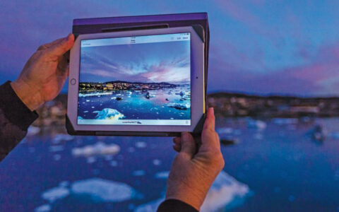 Svalbard, Iceland & Greenland's East Coast traveler holds iPad out to photograph icebergs under a blue & purple sunset.