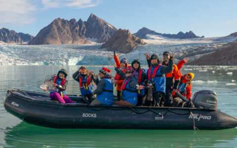 Zodiac dinghy with Svalbard travelers in colorful cold-weather outerwear sits in calm sea beside an icy blue glacier in the sun.