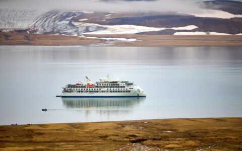 Small white expedition ship sits in glassy fjord while Zodiac cruises nearby with golden tundra on 1 shore & snow on the other.