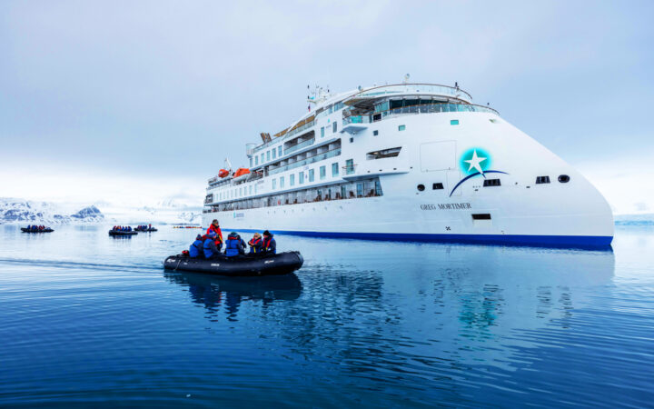 A small Zodiac with Svalbard Odyssey travelers cruising around a small, modern, white cruise ship in the Arctic.