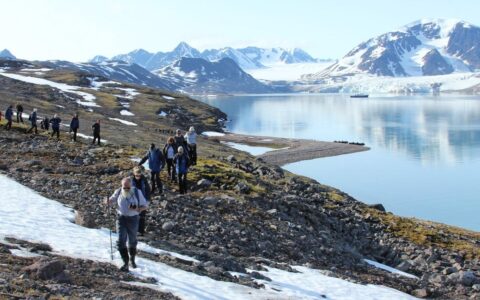 A group of travelers hike toward the camera on a rocky shoreline with the ocean, a ship and mountains behind them in the distance.