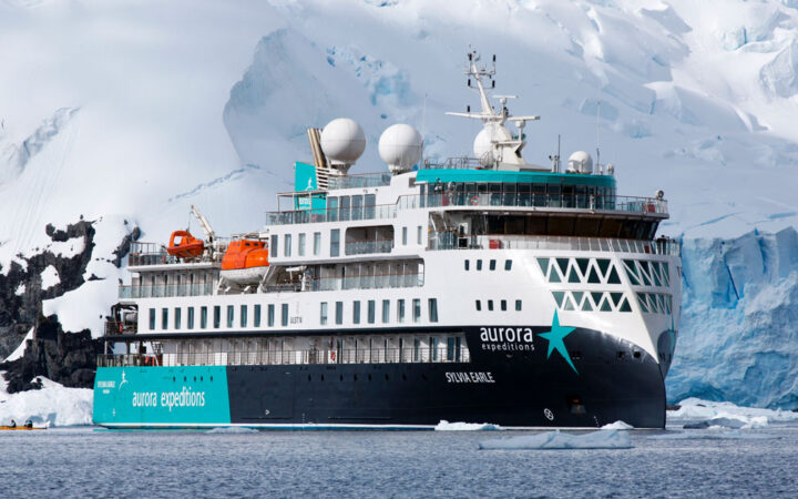 Sylvia Earle ship sitting in calm water, with blue & teal exterior with star logo, 5 white upper decks & 2 orange life boats.