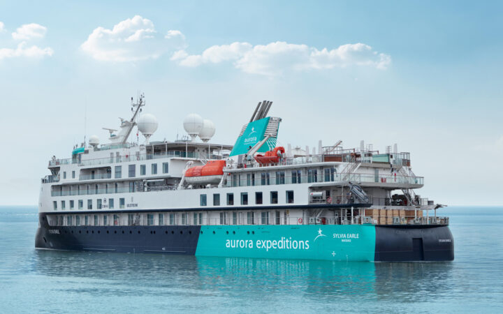 Sylvia Earle ship sitting in calm water, with blue & teal exterior with star logo, 5 white upper decks & 2 orange life boats.