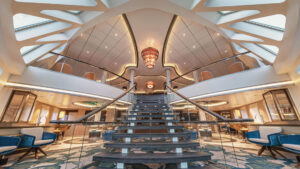 Library on Sylvia Earle ship with central staircase, blue & white carpet, glass rectangle ceiling tiles, chandelier & 2 floors.