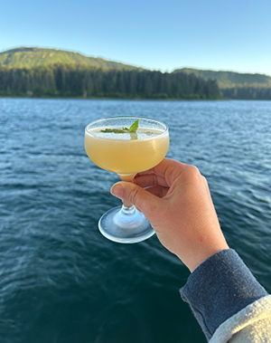 From a ships deck, an arm and hand extend out holding a colorful yellow cocktail with water and forested alaska landscape behind.