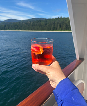 From a ships deck, an arm and hand extend out holding a colorful red cocktail with water and forested alaska landscape behind.