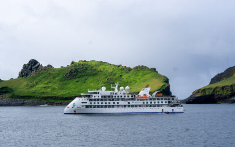 Small white expedition ship with pointed bow sits in calm water by green cliffs on a cloudy day of the Wild Scotland cruise.