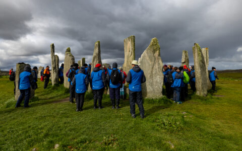 Small group of Wild Scotland cruise travelers in blue jackets walk among primitive tall stone structures in a grassy field.