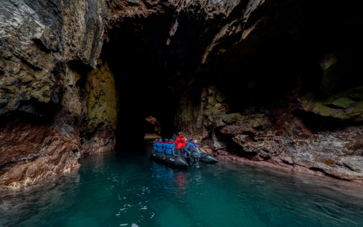 Wild Scotland cruise travelers in blue jackets with guide in red parka ride a Zodiac boat into a cave in turquoise water.