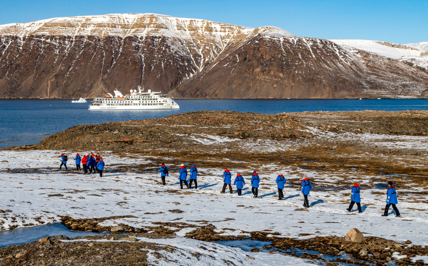 Travelers trecking through the snowy and grassy landscapes off a small stream with their small ship cruise off in the distance