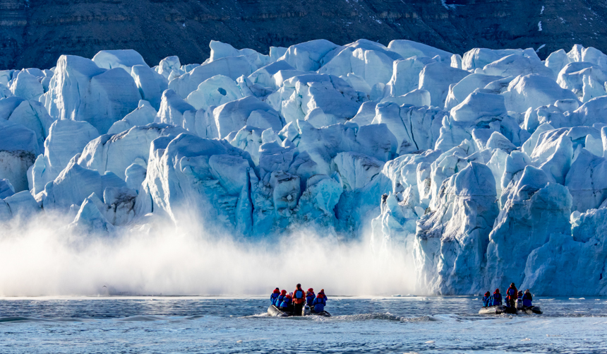 Massive glaciers calving into the ocean as guests watch from nearby zodiacs in awe on a bright sunny day