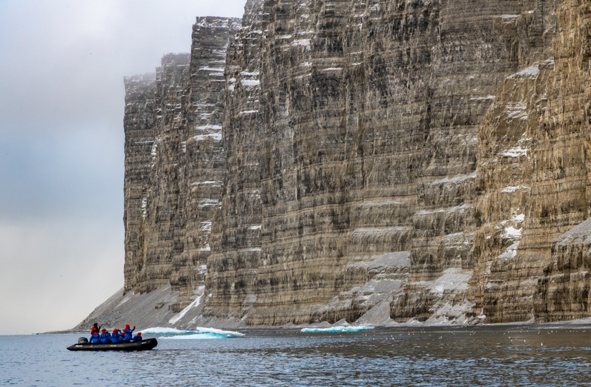 Guests aboard a small cruise ship explore in a Zodiac off of steep tall jagged cliffs with a light dusting of snow