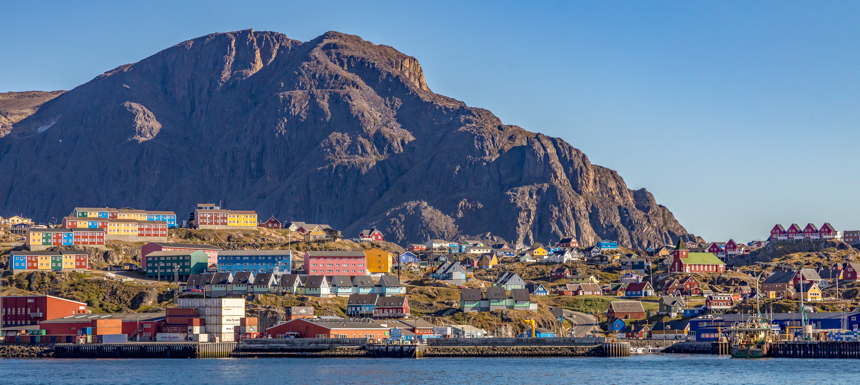 View off of small ship looking inland to the colorful buildings off Greenland with tall mountains towering behind on a sunny day