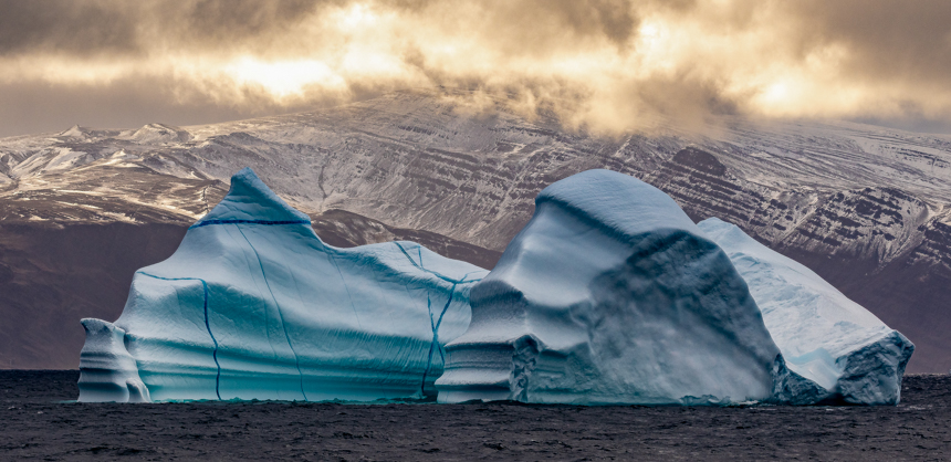 A beautiful white and blue iceberg floating in the middle of the ocean in front of dark grey and brown mountains with bright clouds above