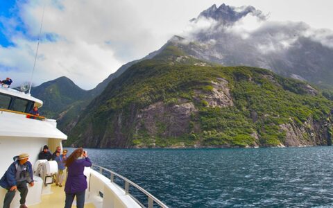Several expedition cruise guests on the bow of their ship in New Zealand photographing the tall green coastal mountains with their peaks in the clouds.