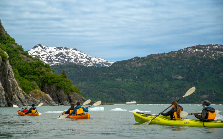 Kayakers in yellow lifevests, paddling through the bright tall mountains in alaska with snow off in the distance