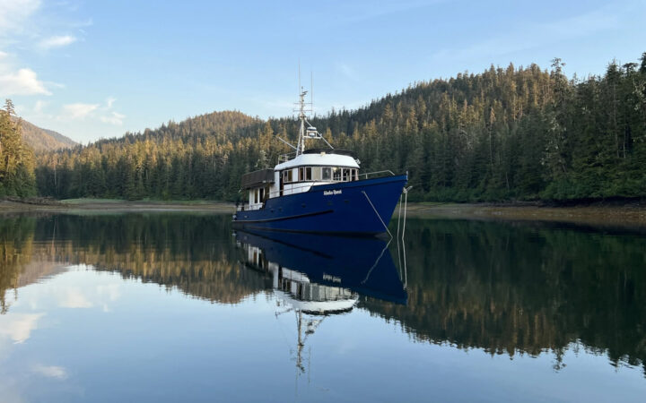 Exterior of Alaska Quest small charter yacht with dark blue hull, white pilothouse & upper deck, on anchor in glassy, secluded cove.