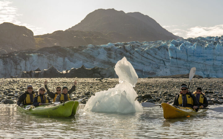 In Alaska a group of kayakers float in green and yellow kayaks next to a white iceberg and in front of an icy teal glacier.