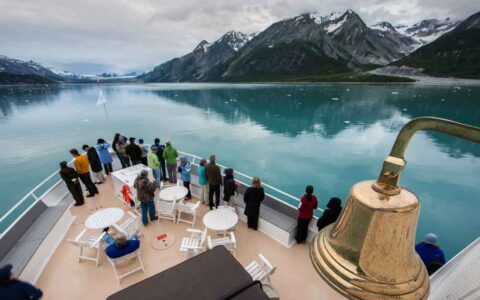 Travelers stand on bow of small ship with gold bell & white tables looking onto teal water with snowcapped peaks behind.