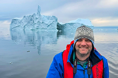AdventureSmith's Andrew Browning wears beanie and royal blue parka behind him floats a massive iceberg.