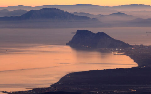 A scenic photograph of the Rock of Gibraltar at sunrise or sunset, with the rock formation silhouetted against an orange and purple sky and reflected in the calm water of the Strait of Gibraltar.