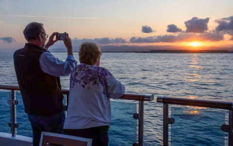A person on a cruise ship deck holds up a phone to take a picture of a sunset over the ocean. Another person stands next to them, also looking at the sunset.