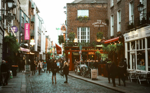 Travelers on a British Isles & France cruise stand on cobblestone streets among bars, restaurants & shops in evening light.