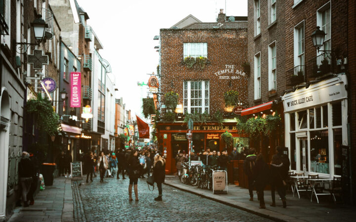 Travelers on a British Isles & France cruise stand on cobblestone streets among bars, restaurants & shops in evening light.