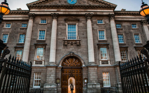 British Isles cruise traveler stands in doorway of large stone Trinity College building, by iron gates, at dusk.