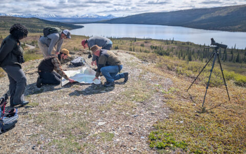 Small group of Alaska travelers kneel over a map beside a telescope on a summit overlooking a lake & snowy peaks in the sun.