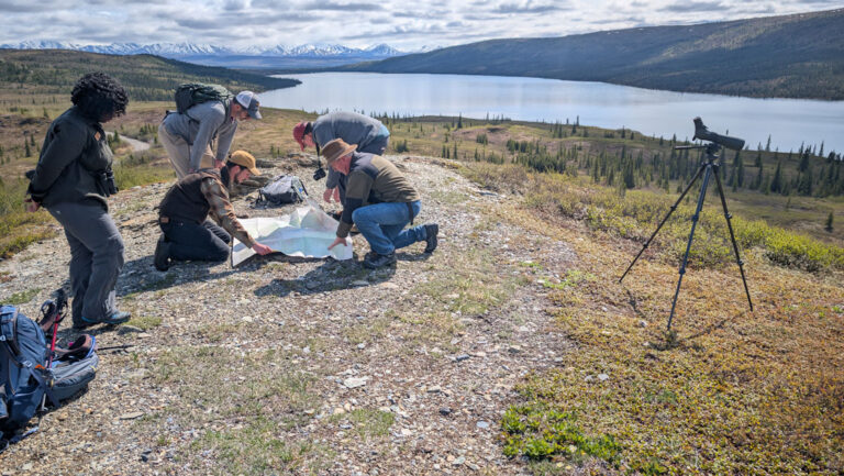 Small group of Alaska travelers kneel over a map beside a telescope on a summit overlooking a lake & snowy peaks in the sun.
