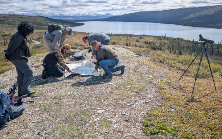 Small group of Alaska travelers kneel over a map beside a telescope on a summit overlooking a lake & snowy peaks in the sun.