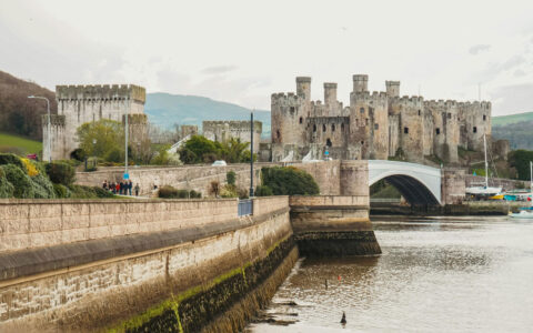 A river with walled banks leads to a large beige castle set among green hills in Conwy, Wales.