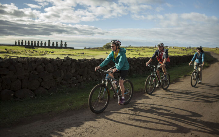 Explora travelers on Easter Island tours peddle mountain bikes on flat dirt road by bright green fields with moai stone statues.
