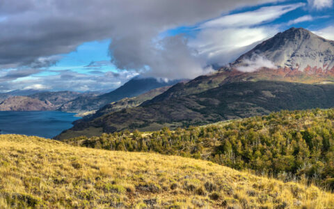 Landscape of grassy hills & volcanic mountains beside a bright blue lake under a partly cloudy sky in Patagonia National Park.