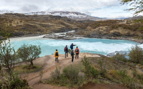 4 Explora Patagonia National Park travelers stand beside a large turquoise river as a guide points out flora & fauna.