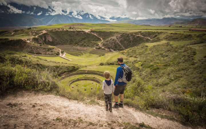 Man & child stand atop a grassy field overlooking a large crater with grassy circles in Sacred Valley, Peru.