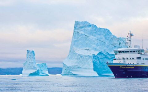 Nat Geo Explorer small ship with dark blue hull & white upper decks cruises by a large iceberg taller than the ship on the Fabled Lands of The North: Greenland to Newfoundland small ship cruise.