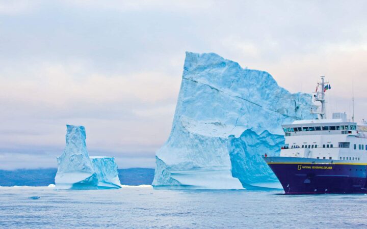 Nat Geo Explorer small ship with dark blue hull & white upper decks cruises by a large iceberg taller than the ship on the Fabled Lands of The North: Greenland to Newfoundland small ship cruise.