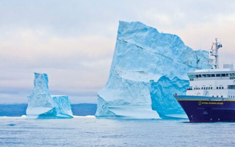 Nat Geo Explorer small ship with dark blue hull & white upper decks cruises by a large iceberg taller than the ship on the Fabled Lands of The North: Greenland to Newfoundland small ship cruise.