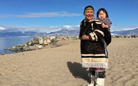 Inuit woman with small child on her back stands on a sandy beach during the Fabled Lands of The North: Greenland to Newfoundland voyage.