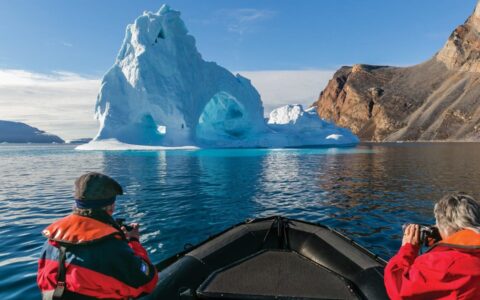 Travelers sit in a Zodiac & photograph a large iceberg beside beige rock on a cruise from Greenland to Canada.